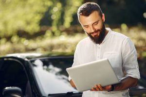 Jeune homme barbu et souriant avec un ordinateur dans les mains recherchant une assurance auto. Voiture noire au deuxième plan.